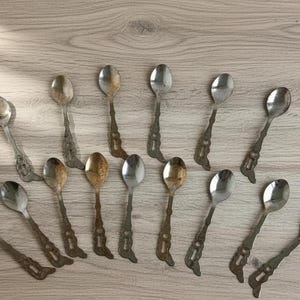 Puede incluir: Un surtido de cucharas de plata vintage con diseños ornamentados en los mangos. Las cucharas, en tonos plateados y dorados, están dispuestas sobre una superficie de madera clara. Colección de cubiertos antiguos.