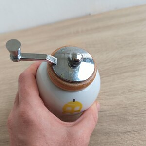 Op de afbeelding: Een witte keramische pepermolen met een houten basis en een zilverkleurige metalen slinger. De molen heeft een klein geel appelontwerp aan de zijkant. De slinger is bevestigd aan een zilverkleurige metalen bovenkant.
