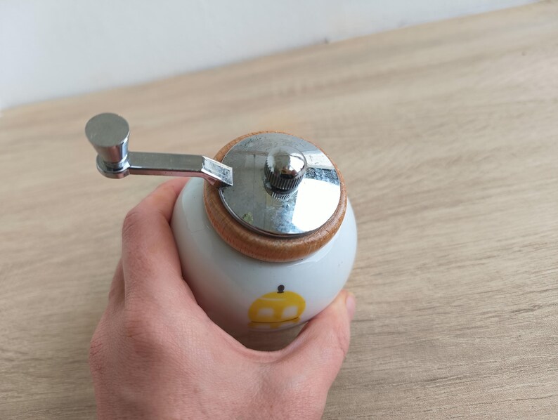 Op de afbeelding: Een witte keramische pepermolen met een houten basis en een zilverkleurige metalen slinger. De molen heeft een klein geel appelontwerp aan de zijkant. De slinger is bevestigd aan een zilverkleurige metalen bovenkant.