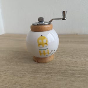 A small, round vintage-style pepper mill with a white porcelain body and a light wooden top. The mill features a subtle yellow geometric graphic on its side and has an exposed metal grinding mechanism at the top. The image is a close-up, showcasing the mill's design and internal structure, resting on a rustic farmhouse wooden table.