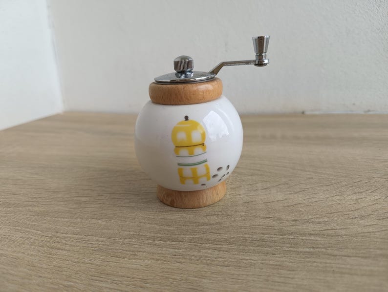 A small, round vintage-style pepper mill with a white porcelain body and a light wooden top. The mill features a subtle yellow geometric graphic on its side and has an exposed metal grinding mechanism at the top. The image is a close-up, showcasing the mill's design and internal structure, resting on a rustic farmhouse wooden table.