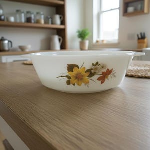 May include: A white, oval-shaped serving bowl with a floral design featuring yellow, white, and red flowers. The bowl has two small handles and sits on a light-colored wooden table. Kitchenware.