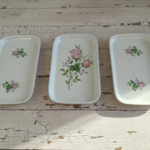 Op de afbeelding: Drie rechthoekige witte keramische serveerschalen. Elke schaal heeft een bloemenontwerp met roze rozen en groene bladeren. De schalen zijn gerangschikt op een wit houten oppervlak met een verweerde uitstraling.