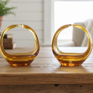 Könnte beinhalten: Zwei bernsteinfarbene Glasvasen mit einem einzigartigen Griffdesign. Die Vasen haben eine abgerundete Form mit flachem Boden und einem kreisförmigen Griff, der sich nach oben wölbt. Sie stehen auf einem Holztisch, mit unscharfem Hintergrund.