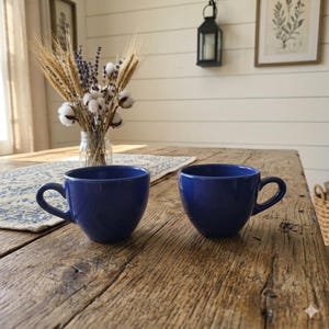 Puede incluir: Dos tazas de café de cerámica azul cobalto con asas curvas sobre una mesa de madera rústica. Las tazas están vacías y tienen un acabado brillante. La mesa está decorada con un arreglo floral y un camino de mesa estampado.