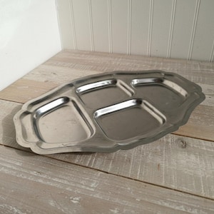 May include: A vintage silver-toned divided serving tray with four compartments. The tray has a scalloped edge and is displayed on a light-colored wooden surface. Ideal for serving appetizers or snacks.