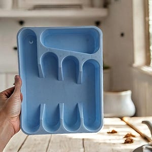 K&ouml;nnte beinhalten: Ein hellblaues Plastik-Bestecktablett mit f&uuml;nf F&auml;chern zum Aufbewahren von Besteck. Das Tablett wird von einer Hand gehalten.