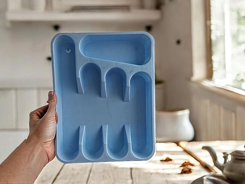 K&ouml;nnte beinhalten: Ein hellblaues Plastik-Bestecktablett mit f&uuml;nf F&auml;chern zum Aufbewahren von Besteck. Das Tablett wird von einer Hand gehalten.