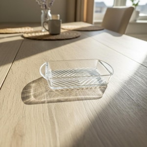 Puede incluir: Una fuente rectangular de vidrio transparente con asas, sobre una mesa de madera clara. La fuente tiene un fondo texturizado y estampado. La luz del sol proyecta sombras sobre la mesa, resaltando la forma y los detalles de la fuente.