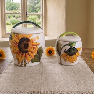 Pode incluir: Dois potes de cerâmica brancos com tampas, decorados com girassóis amarelos e detalhes verdes. Um pote é maior com uma alça verde, o outro é menor. Os potes estão sobre um caminho de mesa de linho bege.