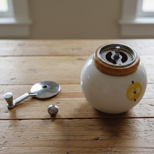 A small, round vintage-style pepper mill with a white porcelain body and a light wooden top. The mill features a subtle yellow geometric graphic on its side and has an exposed metal grinding mechanism at the top. The image is a close-up, showcasing the mill's design and internal structure, resting on a rustic farmhouse wooden table.