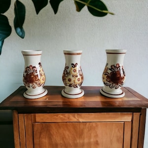 May include: Three white ceramic vases with brown and yellow floral designs. The vases are arranged on a wooden surface.