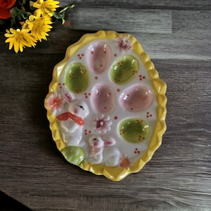May include: A white ceramic egg plate with a yellow rim and a scalloped edge. The plate has six egg-shaped indentations and is decorated with bunnies, flowers, and green and pink eggs.