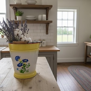 Könnte beinhalten: Ein gelb-weißer Emaille-Eimer mit blauen Blumenmustern enthält Lavendel- und Baumwollstiele. Der Eimer steht auf einem Holztisch in einer hellen Küche mit weißen U-Bahn-Fliesen und Holzregalen.