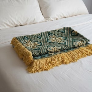 Peut inclure: Une couverture pliée, ornée d'un motif géométrique vert et beige et de franges jaunes, est posée sur un lit blanc. Le motif de la couverture comprend des formes en étoile. Le lit est composé d'oreillers blancs et d'un drap blanc.