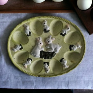May include: A green ceramic egg plate with 12 individual egg cups and bunny rabbit figurines. The plate has a speckled green glaze and a yellow rim.