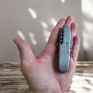 Può includere: Un coltellino svizzero multifunzione in argento con una varietà di strumenti, tra cui una lama, un cavatappi e un cacciavite. Il coltellino è chiuso e tenuto in una mano.