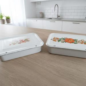 May include: Two white rectangular enamelware containers with lids. Each lid features a floral design in orange, yellow, and green. The containers sit on a light wood surface, with a kitchen in the background.