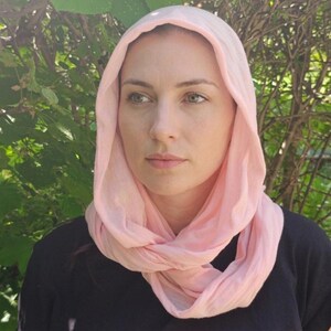 May include: A light pink scarf draped around the head and neck. The scarf is made of a soft, lightweight fabric. The person is wearing a black shirt. The background is a lush green bush.