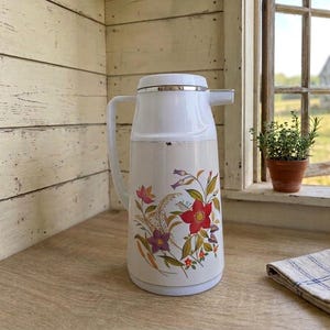 Vintage cream-colored thermos jug with a vibrant retro floral print featuring a red flower, purple blossoms, and wheat. The insulated carafe has a white plastic handle, lid, and pouring spout, accented by a shiny chrome metal trim. Inside, a reflective silver glass vacuum flask is visible.