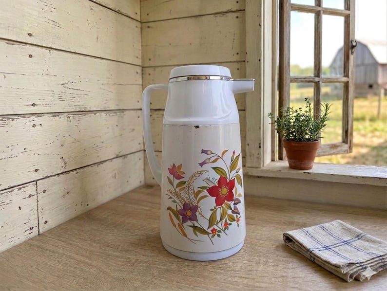 Vintage cream-colored thermos jug with a vibrant retro floral print featuring a red flower, purple blossoms, and wheat. The insulated carafe has a white plastic handle, lid, and pouring spout, accented by a shiny chrome metal trim. Inside, a reflective silver glass vacuum flask is visible.