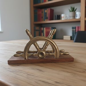 Op de afbeelding: Een messing scheepsrad-brievenweger op een houten voet. Het nautische bureauaccessoire is goudkleurig en staat op een lichtgekleurde houten tafel. De achtergrond omvat een boekenkast en een raam.