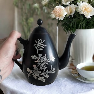 Puede incluir: Una tetera negra con un diseño floral blanco. La tetera tiene un cuerpo redondeado y un pico en el lateral. La tetera está sobre una superficie blanca.