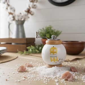 A small, round vintage-style pepper mill with a white porcelain body and a light wooden top. The mill features a subtle yellow geometric graphic on its side and has an exposed metal grinding mechanism at the top. The image is a close-up, showcasing the mill's design and internal structure, resting on a rustic farmhouse wooden table.