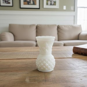 Peut inclure: Un petit vase blanc de style vintage avec une surface texturée et à motifs. Le vase est posé sur une table en bois. Le vase a une base ronde et un dessus évasé. L'arrière-plan comprend un canapé et un livre.