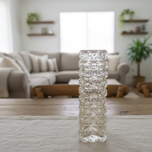 Può includere: Un vaso di vetro trasparente con un motivo geometrico strutturato. Il vaso cilindrico è posto su un tavolo di legno. Lo sfondo mostra un soggiorno con un divano, sedie di legno e una finestra.