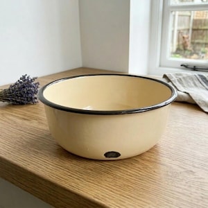 May include: Cream-colored enamel bowl with a black rim. The bowl sits on a wooden surface, with a small black spot on the side. A bundle of lavender and a striped linen towel are in the background.