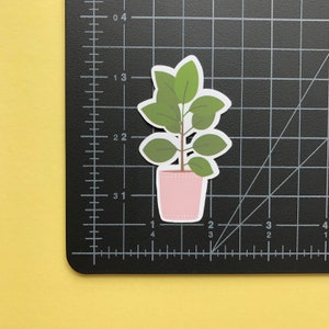 May include: A die-cut sticker of a potted plant with green leaves and a light pink pot, positioned on a black cutting mat with white grid lines. The sticker measures approximately 2 inches in height and 1.25 inches in width as shown.