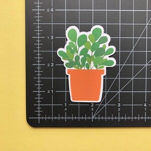 May include: A die-cut sticker of a potted plant with rounded green leaves in a solid orange pot. The sticker is placed on a black cutting mat with white grid lines, showing the plant is approximately 2.5 inches tall and 2 inches wide.