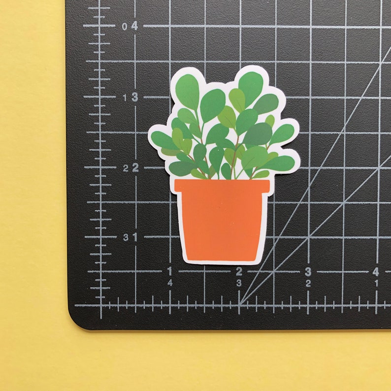 May include: A die-cut sticker of a potted plant with rounded green leaves in a solid orange pot. The sticker is placed on a black cutting mat with white grid lines, showing the plant is approximately 2.5 inches tall and 2 inches wide.