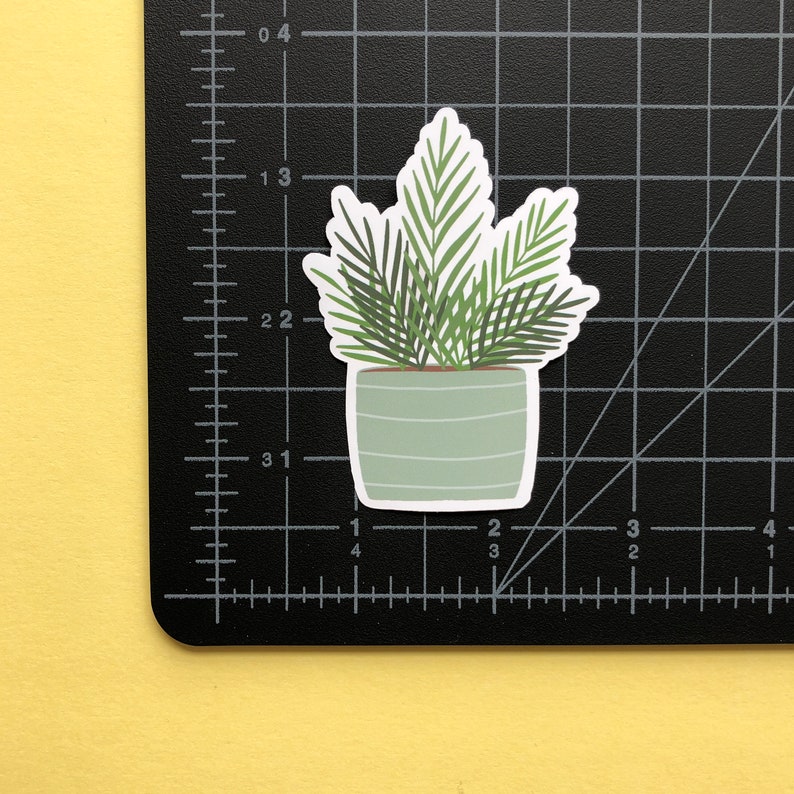 May include: A die-cut sticker of a potted palm plant with dark green fronds in a light sage green pot with horizontal white stripes. The sticker is placed on a black cutting mat with white grid lines, showing a height of approximately 2.5 inches.