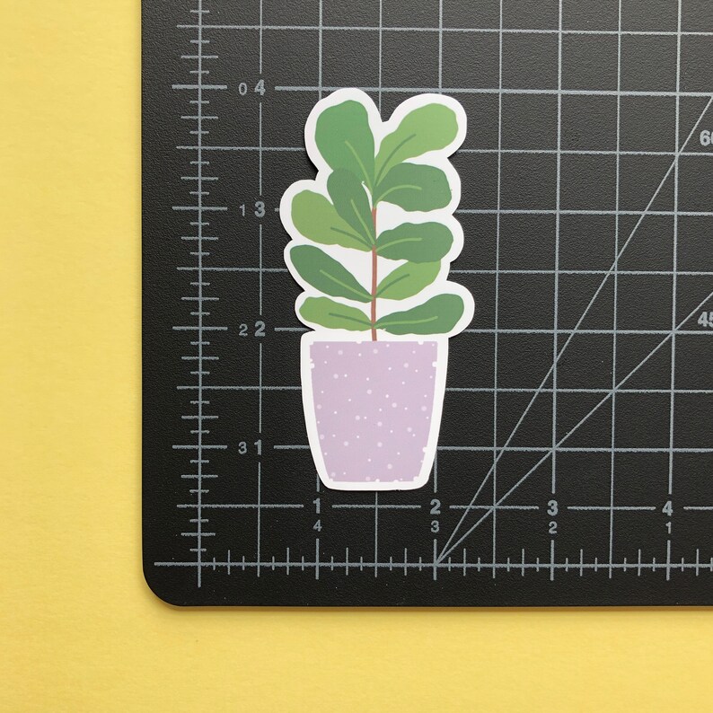 May include: A die-cut sticker of a potted plant with green leaves and a brown stem in a light purple speckled pot. The sticker is placed on a black cutting mat with white grid lines, showing the plant is approximately 3 inches tall.