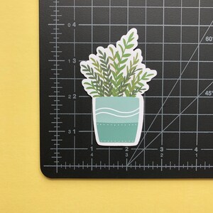 May include: A die-cut sticker of a potted plant with green leaves in a light blue pot featuring white wavy lines and a dotted pattern. The sticker is placed on a black cutting mat with a grid showing measurements in inches, indicating a height of 2 inches.