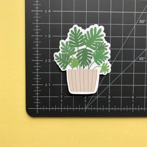 May include: A die-cut sticker of a potted houseplant with green, lobed leaves in a beige, vertically-striped pot. The sticker is placed on a black cutting mat with white grid lines and markings, showing the sticker is approximately 2.5 inches wide.