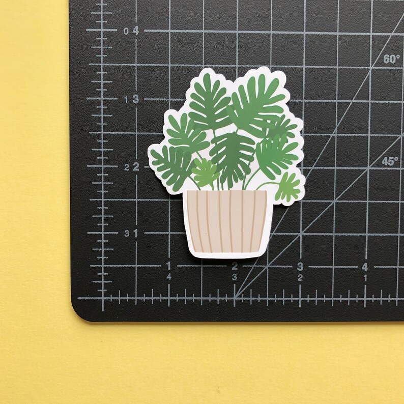 May include: A die-cut sticker of a potted houseplant with green, lobed leaves in a beige, vertically-striped pot. The sticker is placed on a black cutting mat with white grid lines and markings, showing the sticker is approximately 2.5 inches wide.