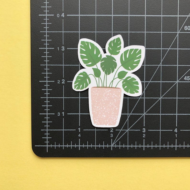 May include: A die-cut sticker of a potted monstera plant with green leaves and a speckled light pink pot. The sticker is placed on a black cutting mat with white grid lines and imperial measurements, showing the plant is approximately 2.5 inches tall.
