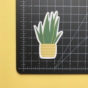 May include: A die-cut sticker of a snake plant in a yellow pot with horizontal stripes. The plant has tall, pointed green leaves with darker green horizontal patterns. The sticker is placed on a black cutting mat with white grid lines in inches.