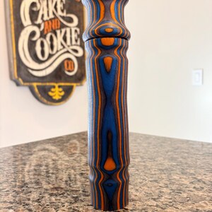 K&ouml;nnte beinhalten: Eine hohe Pfefferm&uuml;hle aus Holz mit blau-orangefarbenem Streifenmuster. Die M&uuml;hle hat einen zylindrischen K&ouml;rper und eine ausgestellte Oberseite. Die Holzmaserung ist sichtbar und erzeugt ein einzigartiges Design. Die M&uuml;hle steht auf einer gesprenkelten Arbeitsplatte.