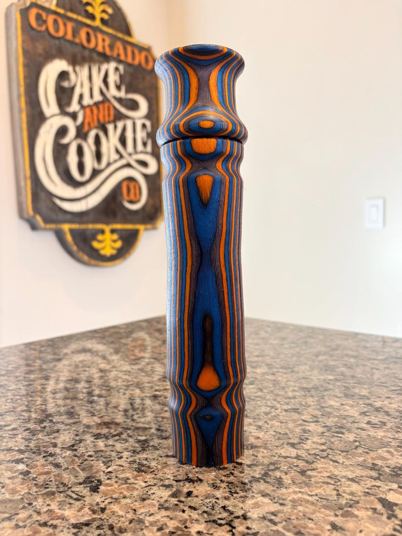 K&ouml;nnte beinhalten: Eine hohe Pfefferm&uuml;hle aus Holz mit blau-orangefarbenem Streifenmuster. Die M&uuml;hle hat einen zylindrischen K&ouml;rper und eine ausgestellte Oberseite. Die Holzmaserung ist sichtbar und erzeugt ein einzigartiges Design. Die M&uuml;hle steht auf einer gesprenkelten Arbeitsplatte.