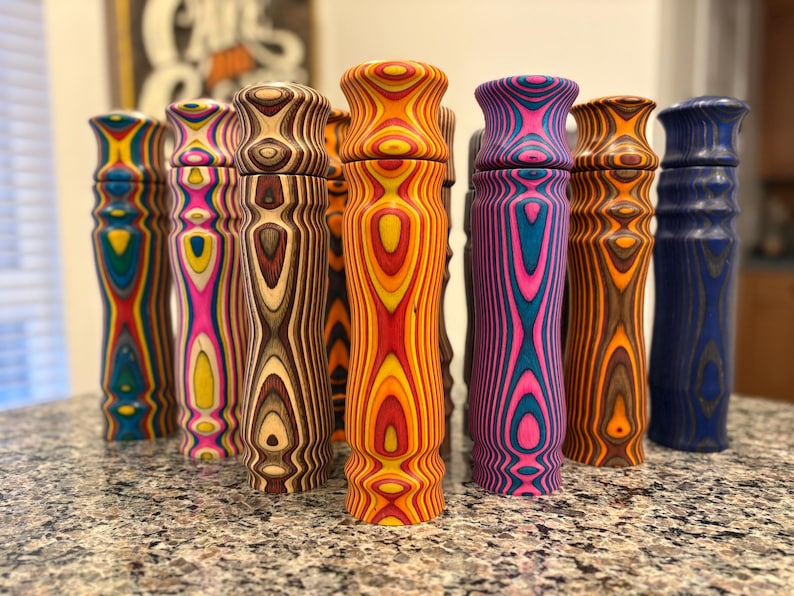 K&ouml;nnte beinhalten: Eine Sammlung farbenfroher Pfefferm&uuml;hlen aus Holz. Die M&uuml;hlen weisen eine Vielzahl lebendiger Farben auf, darunter Blau, Gelb, Rosa und Orange, mit einzigartigen Holzmaserungsmustern. Die M&uuml;hlen sind zylindrisch mit einem ausgestellten Oberteil.