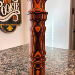 K&ouml;nnte beinhalten: Ein dekorativer Pfefferm&uuml;hle aus Holz mit einem markanten Schichtdesign. Das Holz weist ein auff&auml;lliges Muster aus orangefarbenen, lila und braunen Streifen auf. Die M&uuml;hle steht auf einer gesprenkelten Arbeitsplatte und zeigt ihre detaillierte Verarbeitung.