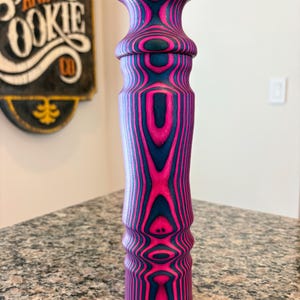K&ouml;nnte beinhalten: Ein dekorativer Holzgegenstand mit einem auff&auml;lligen rosa- und marineblauen Streifenmuster. Der Gegenstand hat einen bauchigen oberen Teil und einen zylindrischen K&ouml;rper, der ein einzigartiges Holzmaserungsdesign zeigt. Er steht auf einer gesprenkelten Arbeitsplatte.
