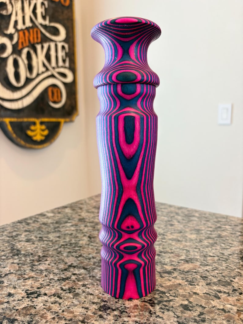 K&ouml;nnte beinhalten: Ein dekorativer Holzgegenstand mit einem auff&auml;lligen rosa- und marineblauen Streifenmuster. Der Gegenstand hat einen bauchigen oberen Teil und einen zylindrischen K&ouml;rper, der ein einzigartiges Holzmaserungsdesign zeigt. Er steht auf einer gesprenkelten Arbeitsplatte.