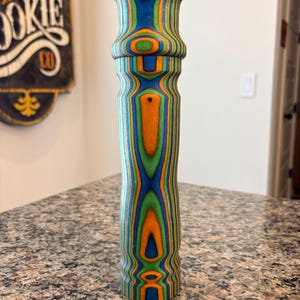 K&ouml;nnte beinhalten: Ein farbenfroher Pfefferm&uuml;hle aus Holz mit einem lebendigen Design. Die M&uuml;hle hat ein geschichtetes Muster aus Blau, Orange und Gr&uuml;n, das einen auff&auml;lligen visuellen Effekt erzeugt. Das Design ist symmetrisch, mit einem ausgestellten oberen und unteren Teil und einem zylindrischen K&ouml;rper.