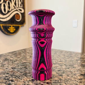 K&ouml;nnte beinhalten: Dekorative Pfefferm&uuml;hle aus Holz mit einem lebendigen rosa-schwarzen Streifenmuster. Die M&uuml;hle hat einen bauchigen Deckel und einen zylindrischen K&ouml;rper, der ein einzigartiges, auff&auml;lliges Design zeigt. Die Holzmaserung ist sichtbar und verleiht dem St&uuml;ck Textur.