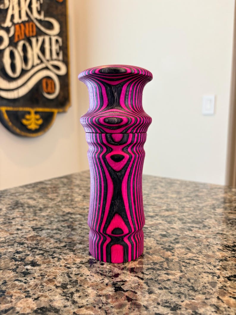 K&ouml;nnte beinhalten: Dekorative Pfefferm&uuml;hle aus Holz mit einem lebendigen rosa-schwarzen Streifenmuster. Die M&uuml;hle hat einen bauchigen Deckel und einen zylindrischen K&ouml;rper, der ein einzigartiges, auff&auml;lliges Design zeigt. Die Holzmaserung ist sichtbar und verleiht dem St&uuml;ck Textur.